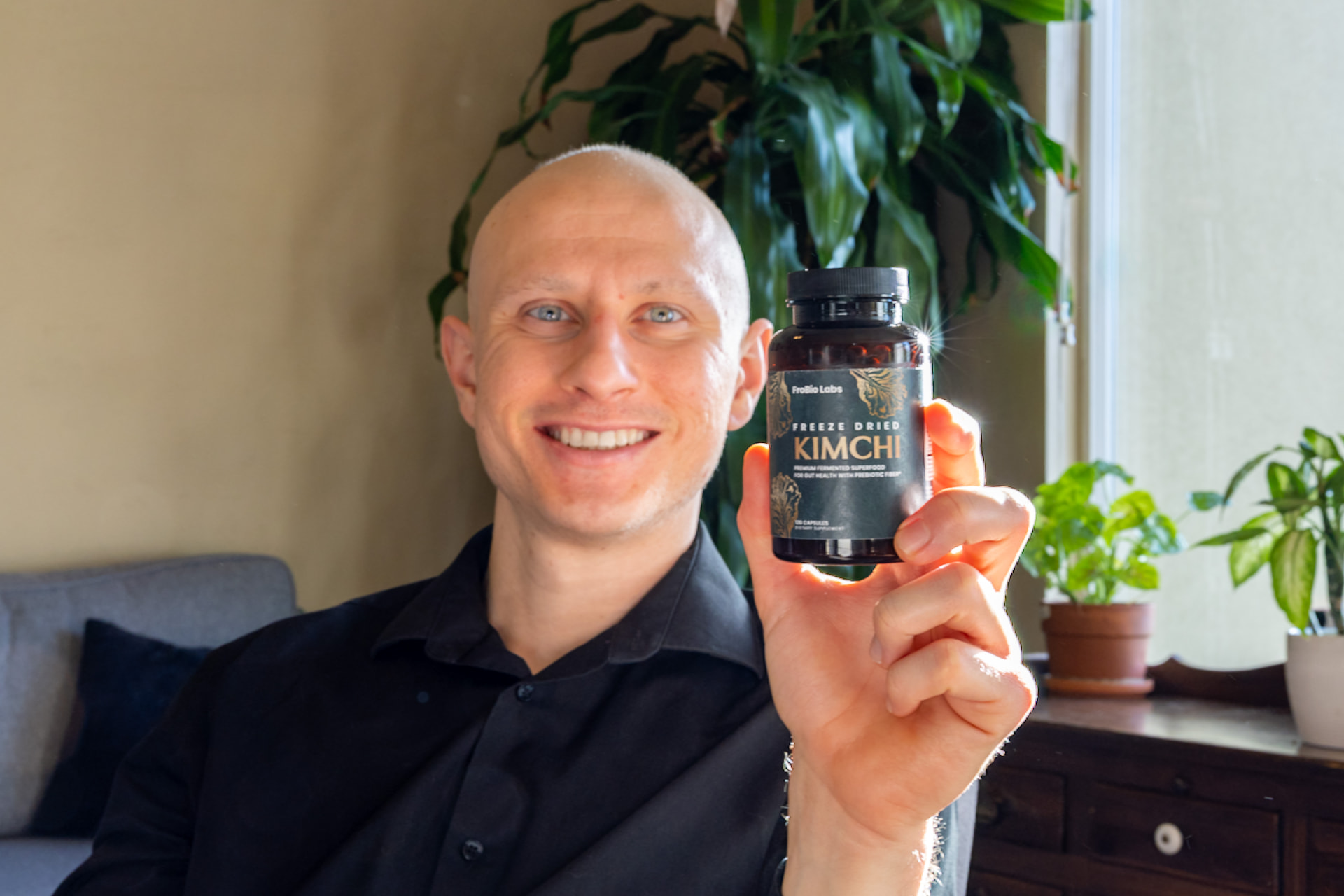 A bottle of FroBio Labs freeze-dried kimchi capsules held up in a sunlit room with houseplants behind